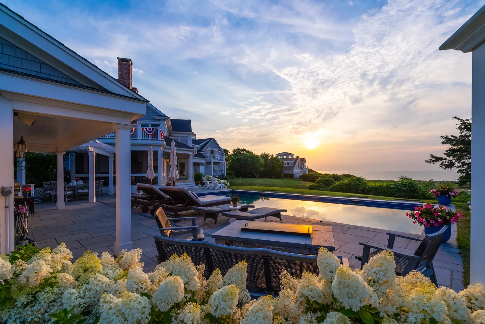 Outdoor living space of a custom Cape Cod home featuring a pool, lounge chairs, stone patio, covered pergola, white hydrangea garden, and a sunset view over the water