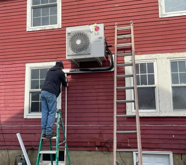 A man standing on a ladder working on an outdoor air conditioning unit mounted on a red house wall