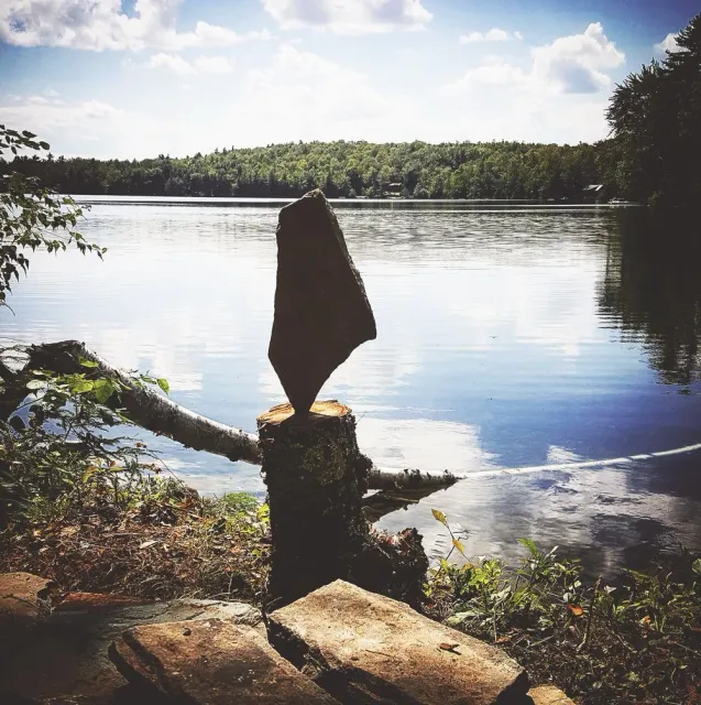 A river scene with a large rock balanced on a tree stump, with a forest in the background and a partly cloudy sky.