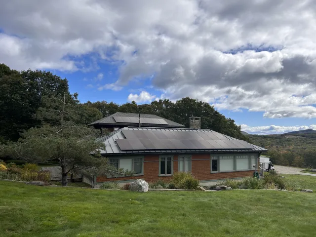 House with solar panels on the roof, surrounded by trees and grassy landscape under a partly cloudy sky.
