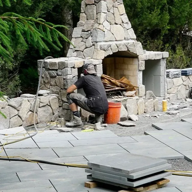 A person working on building or repairing an outdoor stone fireplace with a stone patio in a garden or backyard.