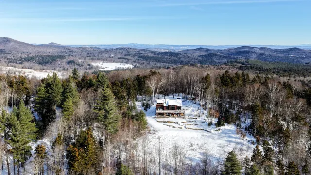 Aerial view of a snow-covered home and clearing within wooded Vermont hills, with distant mountain ridges.