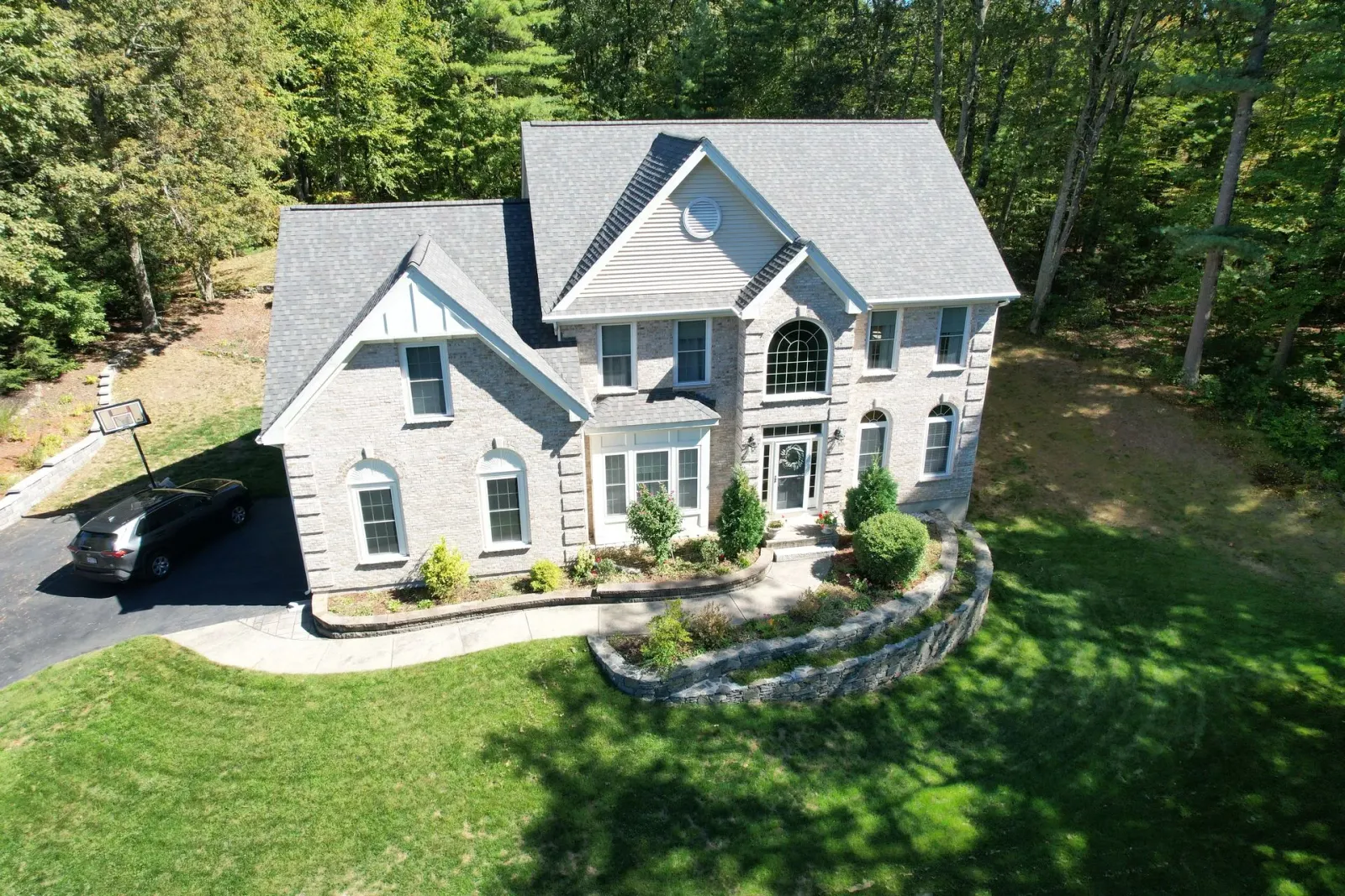 A gray stone house with a dark roof, front porch, and green lawn surrounded by trees.