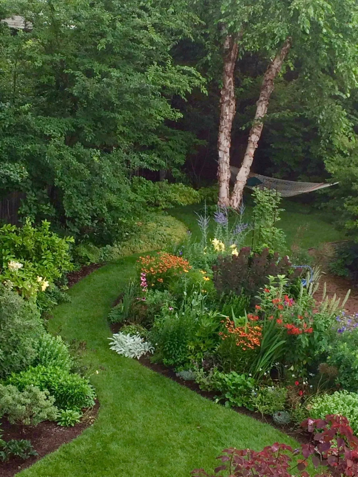 A lush, green garden with a well-maintained lawn, colorful flowers, and leafy trees, including two tall, textured trees and a hammock hanging between two trees in the background.
