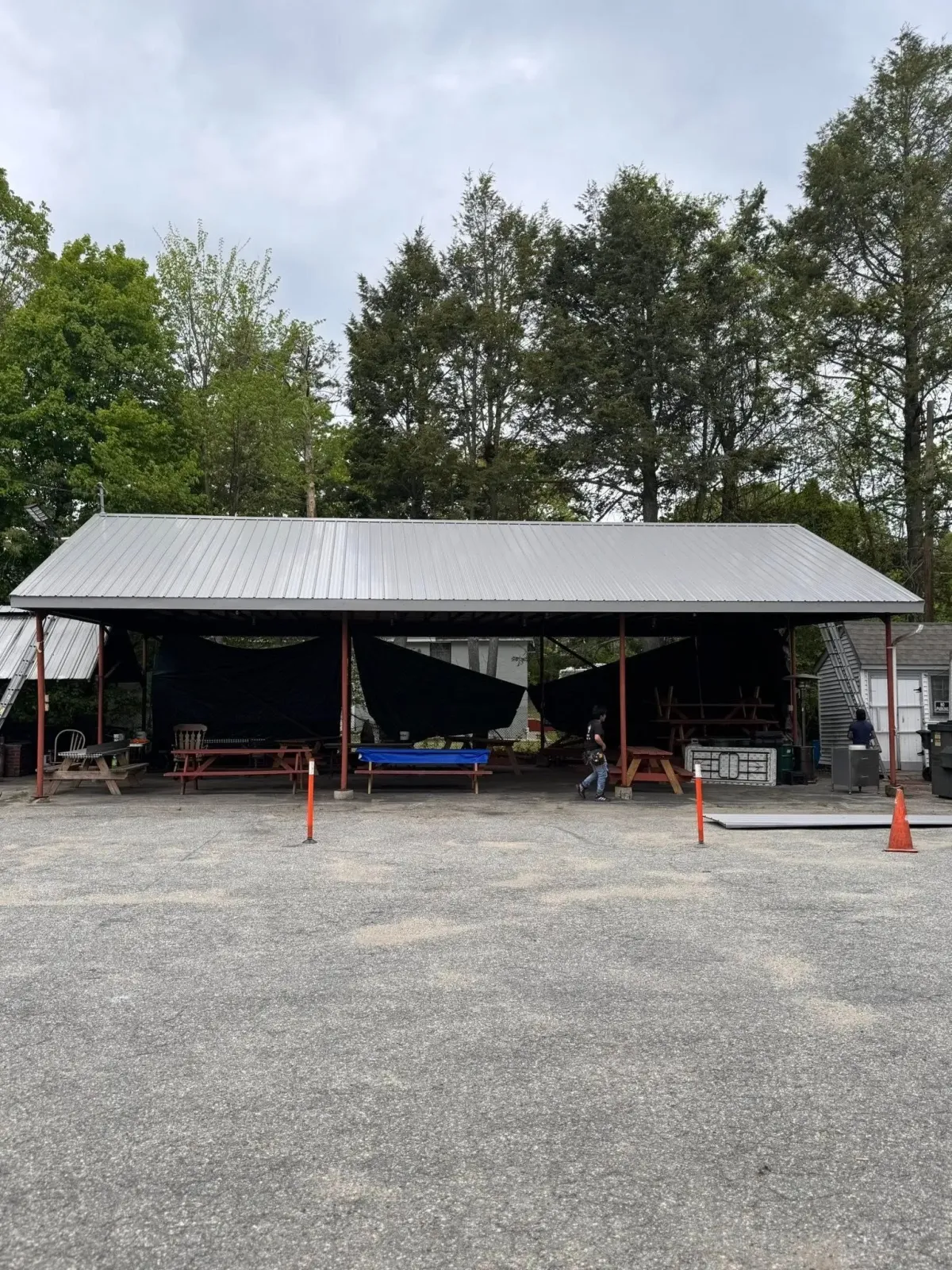 Open-sided metal shed with boats and equipment on a gravel lot, framed by trees and orange cones