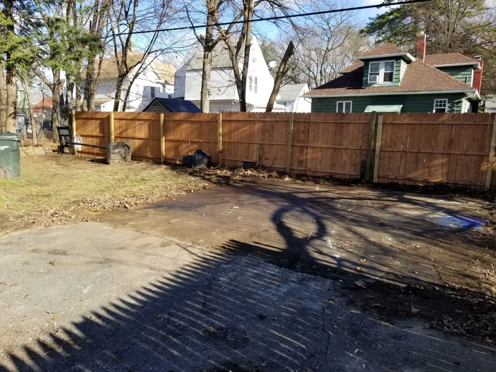 A backyard with a new wooden fence, patches of dirt and pavement, and some outdoor items like trash cans and a chair. There are trees and houses in the background during daytime.