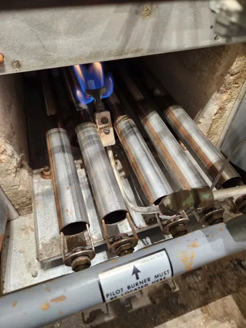 Close-up of a gas heater with five metal tubes, some with visible rust, and a blue flame coming from the pilot burner. There is a label indicating the pilot burner must be located here.