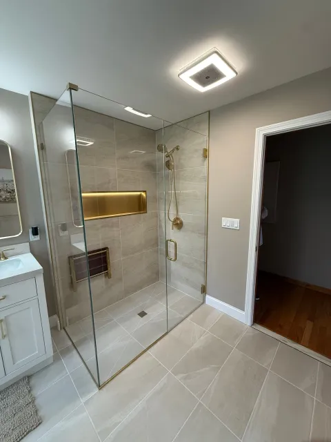 Modern bathroom with glass shower, gold fixtures, beige tile, and open doorway to a dark hall