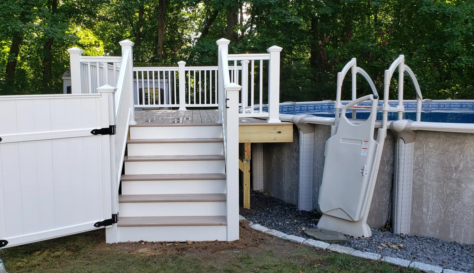 Above-ground pool with a white wooden deck and stairs, surrounded by a white fence, set in a backyard with green trees.