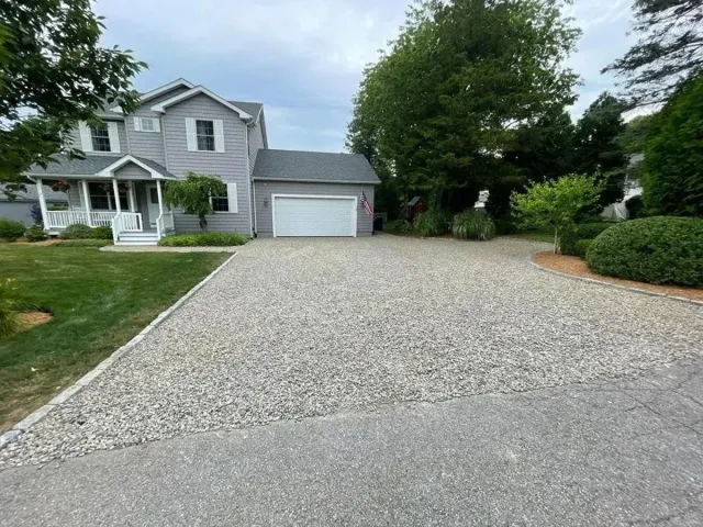 Gravel driveway leading to a gray two-story house with a garage and front porch, surrounded by green trees.