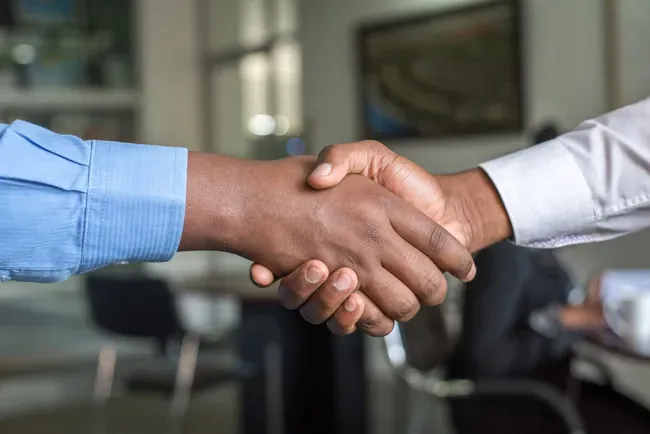 Two people shaking hands in an office, one in a blue shirt and one in a white shirt.