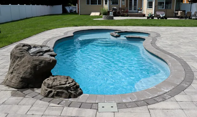 A backyard with a kidney-shaped swimming pool, surrounded by a paved patio and a white fence, with a grassy lawn and a house in the background.