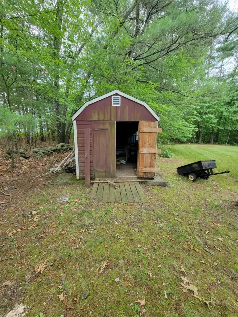 Small wooden shed with a partially open door in a backyard surrounded by green trees and grass, with a black wheelbarrow nearby.