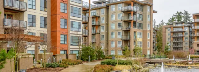 Apartment buildings with varying shades of brown and beige surrounded by landscaping and a water feature. Overcast day.