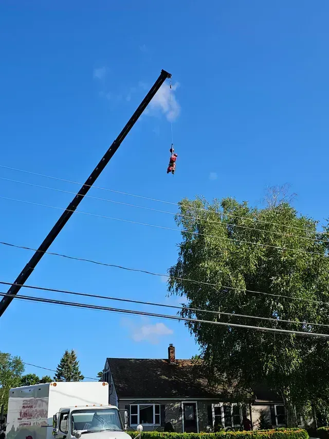 A person in a harness is suspended from a crane, above a house and truck, under a blue sky.