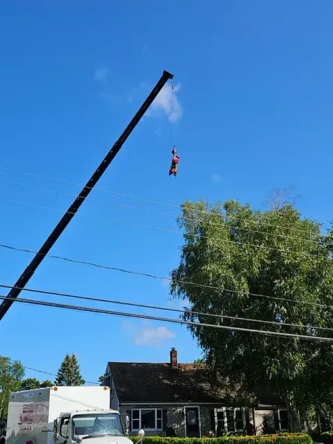 A person in a harness is suspended from a crane, above a house and truck, under a blue sky.