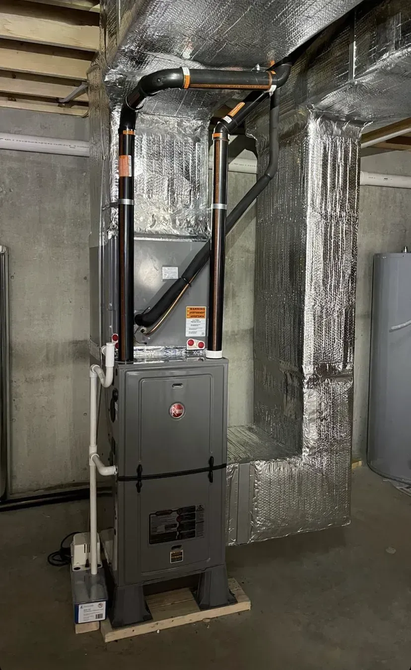 Gray furnace in a basement utility area with insulated ducts and exposed pipes.