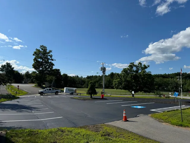 A parking lot with designated handicap parking space, traffic cone, trees, a truck and trailer, utility pole, grassy area, and a field with a basketball hoop in the background under a bright blue sky with scattered clouds.