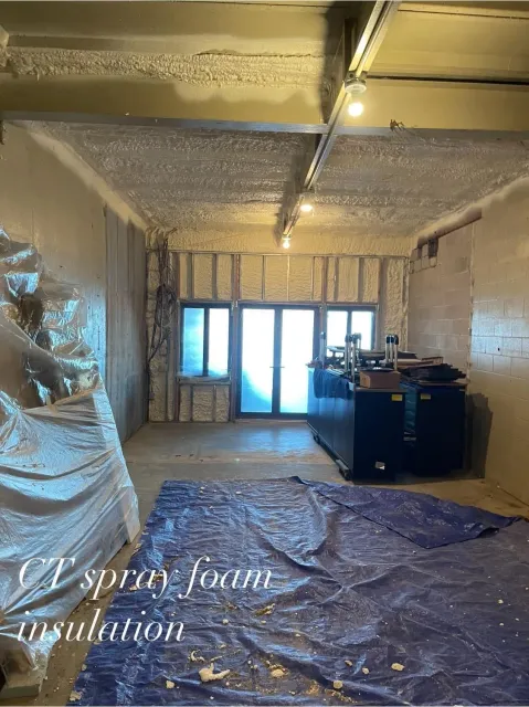 Interior of a room with spray foam insulation on the walls and ceiling, a blue tarp on the floor, and a door with windows.