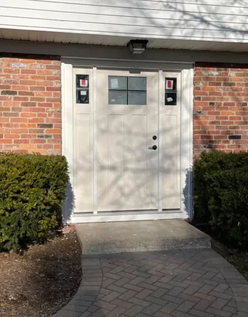 Beige front door with glass panels on a brick building, fronted by a brick path and bushes.
