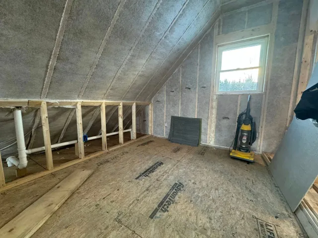 Unfinished attic room with exposed framing, sloped ceiling, small window, and a yellow shop vacuum.