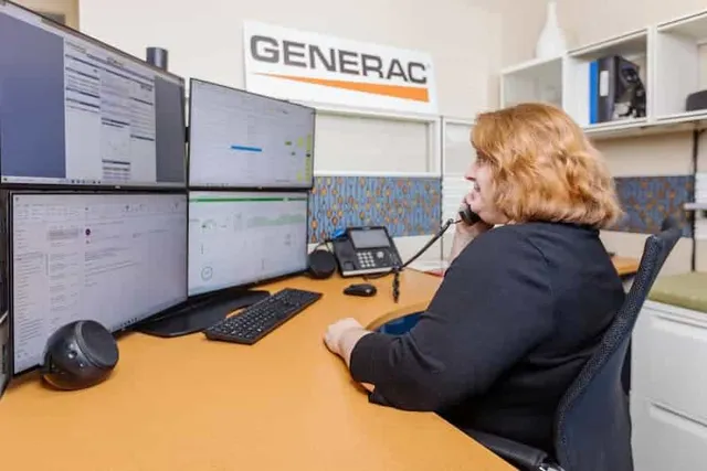 A PE Home Generators employee on the phone at a desk with multiple monitors displaying data, under a Generac sign.