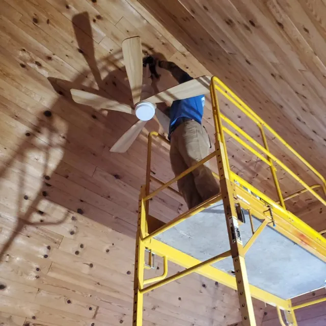 Person on a yellow lift installs a ceiling fan on a wood-paneled ceiling.