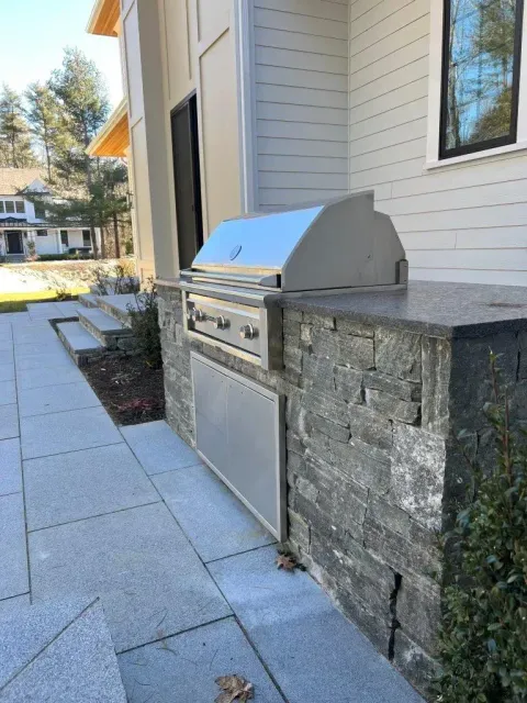 Outdoor stainless steel grill built into a stone counter beside a building walkway