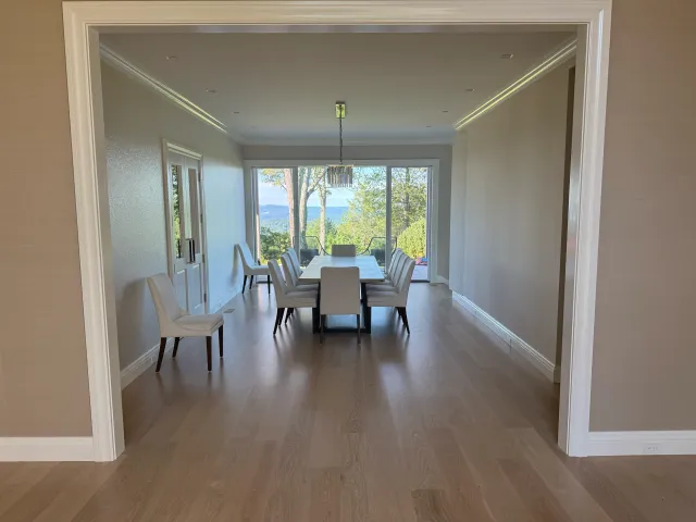 Dining room with wooden floor, white walls, and large sliding glass doors leading to an outdoor view of trees and a body of water.