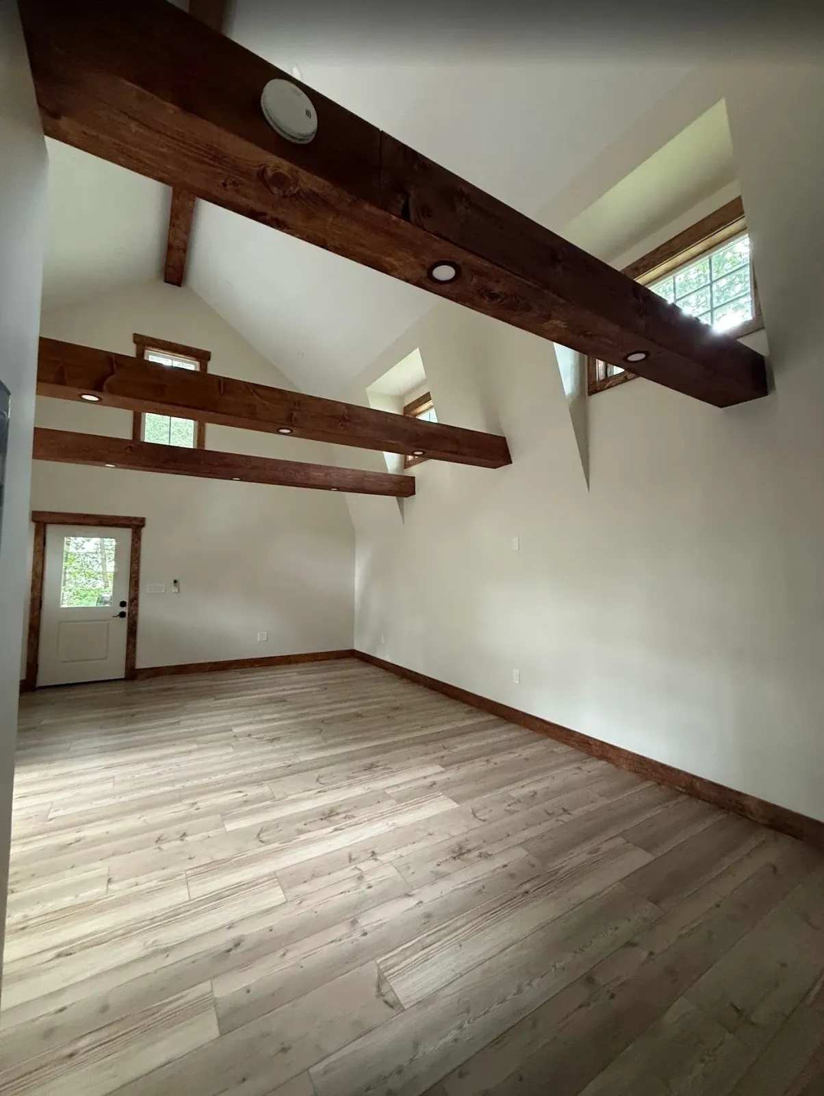 Empty room with light wood floors, white walls, and exposed wooden beams. Door on the left.