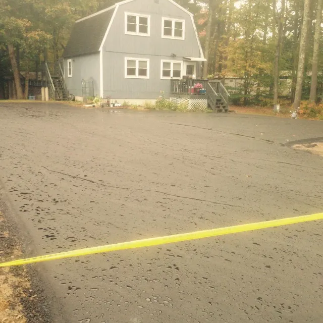 Newly paved driveway in front of a blue house with a gray roof and wooden stairs. Yellow caution tape is across the foreground.