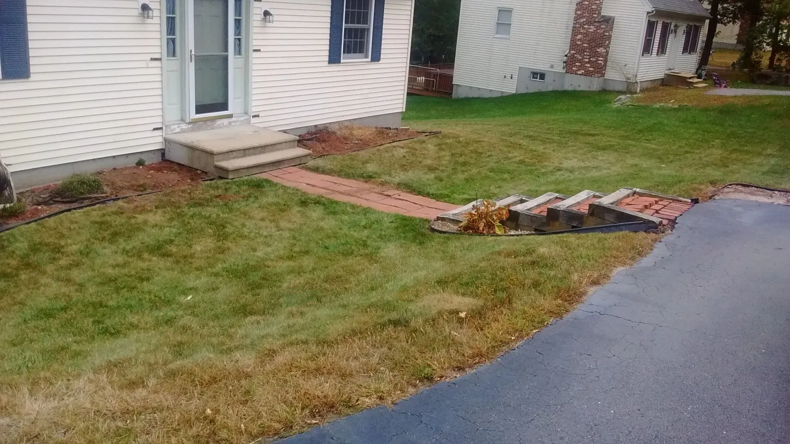 Worn-out concrete steps and a brick walkway in Webster, MA, leading to a house with light siding and blue shutters, surrounded by patchy grass and mulch beds. Project by KRD Landscaping.