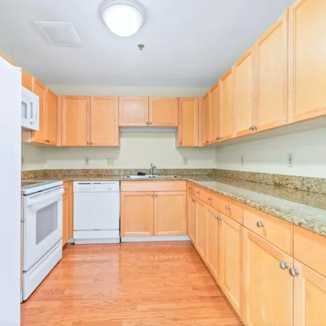 Empty kitchen with light wood cabinets, granite countertops, and white appliances, including a stove and dishwasher.