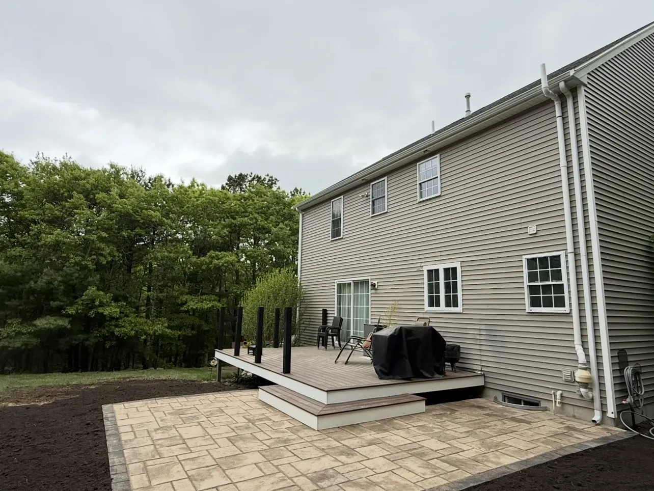 Backyard with deck and paved patio next to a two-story beige house under a cloudy sky.