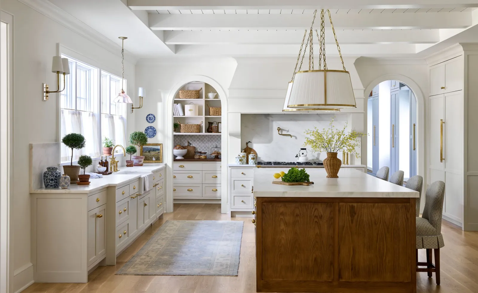 Bright kitchen with white cabinetry and marble countertops, featuring a large wooden island, decorative plants, a chandelier, gold fixtures, and an arched doorway.