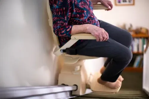 Person seated on a beige stair lift beside a staircase, wearing dark pants and a patterned top.