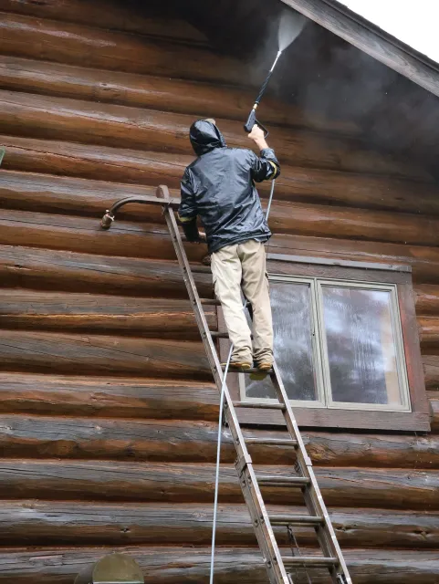 A man is cleaning a log cabin with a high pressure washer.