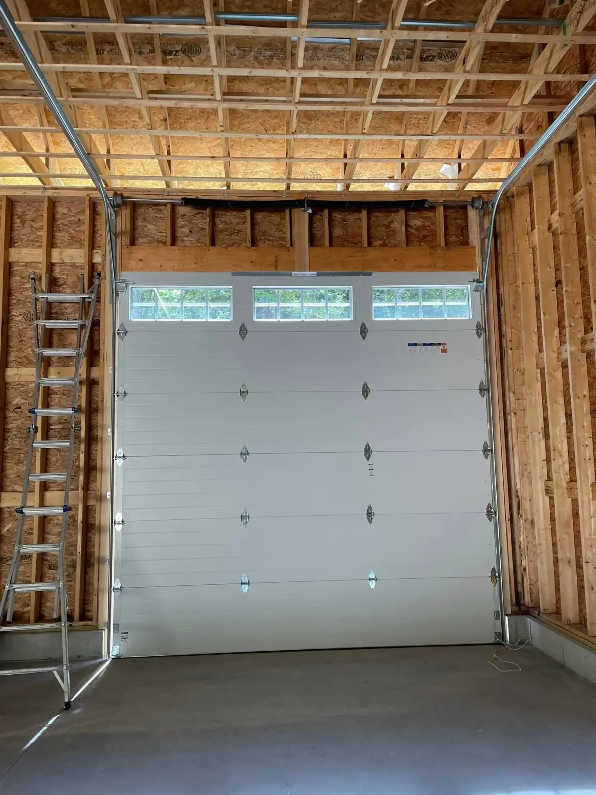 Garage door, white, installed in an unfinished wooden garage with visible framing and ceiling beams.