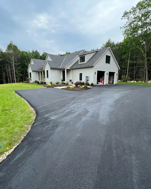 Light gray house with a black asphalt driveway and garage, surrounded by green grass and trees.