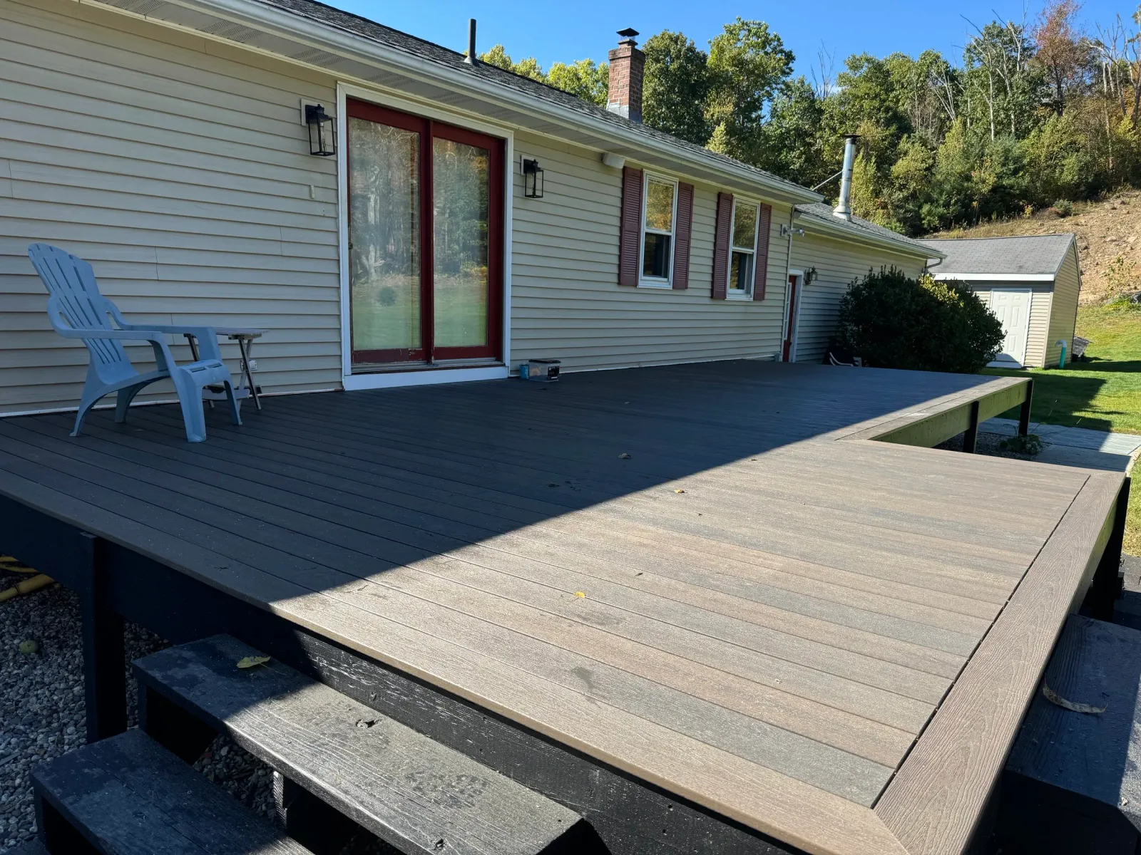Backyard with a wooden deck attached to a house with beige siding, red framed sliding glass door, and two black outdoor wall lights. There's a blue Adirondack chair and small table on the deck, and a small shed in the background with a lawn and hills