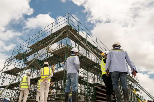 Construction workers in safety vests and hard hats surveying scaffolding under a cloudy sky.