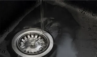 A close-up view of a metal sink drain with water running down into it, creating ripples and reflections on the dark sink surface.