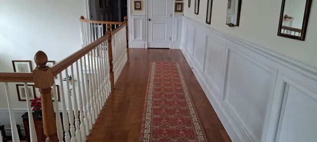 Narrow hallway with white paneled walls, red runner carpet, and wooden railing