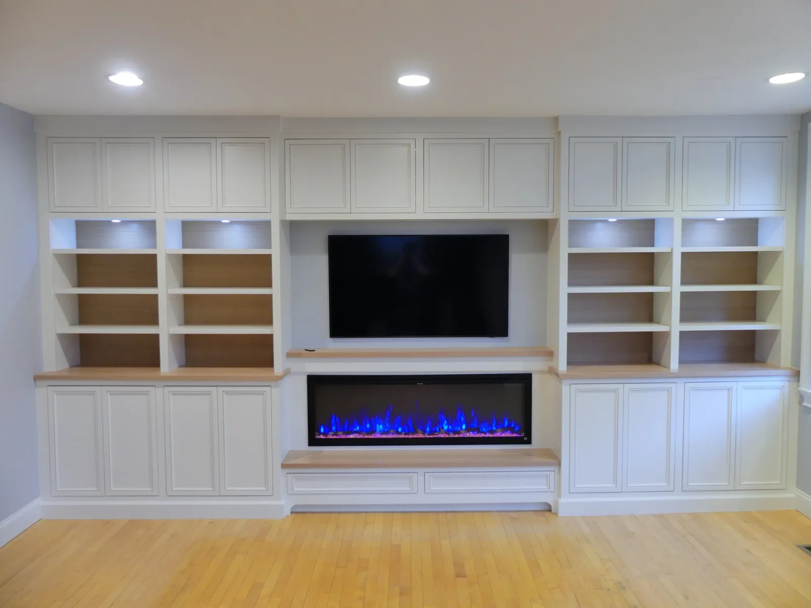 White built-in entertainment center with shelves, cabinets, a wall-mounted television, and an electric fireplace with blue flames on a hardwood floor.