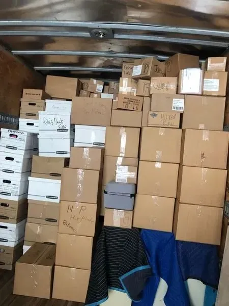Boxes stacked inside a truck, varying sizes and shapes. Some are labeled, against a wooden wall.