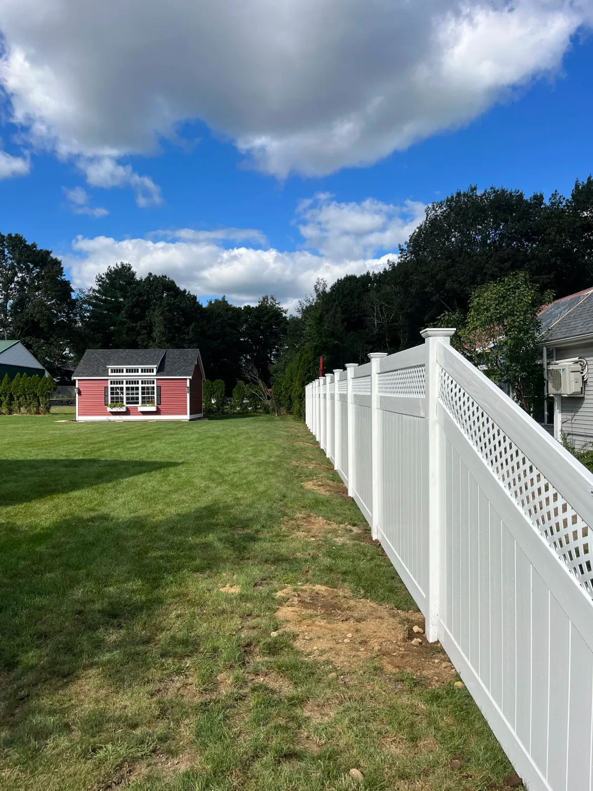 A backyard with green grass, a white vinyl fence, trees, and a pink shed with a black roof. The sky is partly cloudy with blue patches.