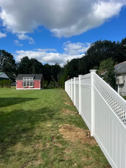 A backyard with green grass, a white vinyl fence, trees, and a pink shed with a black roof. The sky is partly cloudy with blue patches.