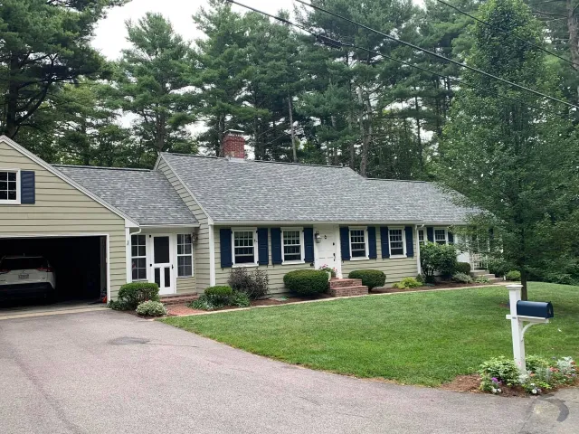 Single-story suburban house with attached garage, gray roof, and green lawn surrounded by trees.