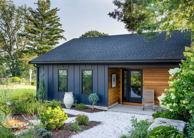 Modern house with dark blue siding, wood accents, lush greenery, and a landscaped garden – Woodland Builders, South Shore MA.