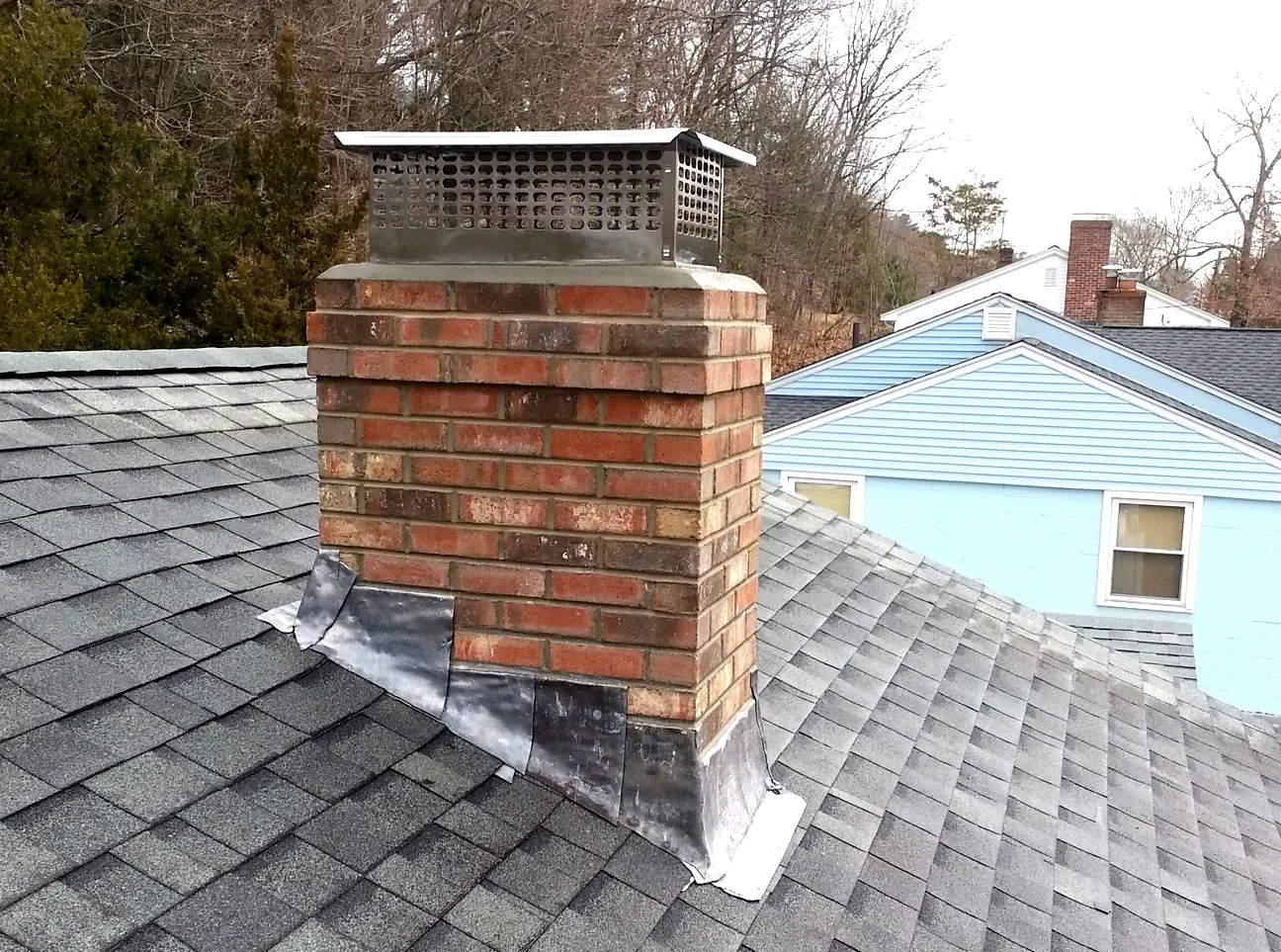 Brick chimney on a shingled roof with a metal cap. Blue house and trees in background.